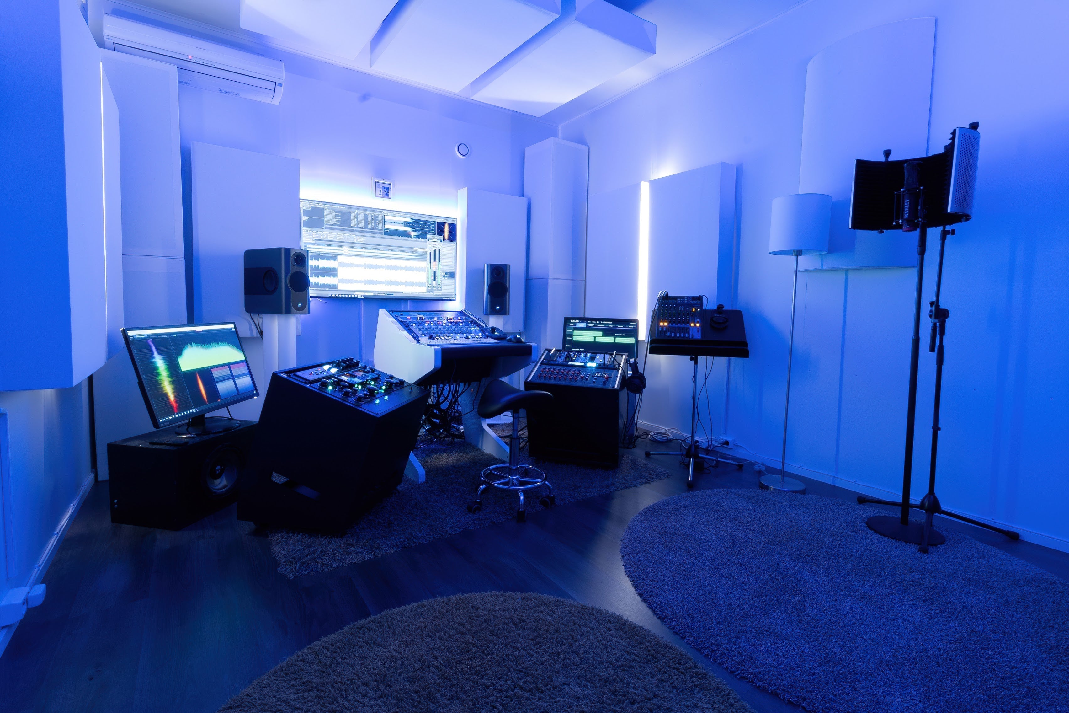 "Recording studio treated with white GIK Acoustics acoustic panels on walls and ceiling clouds, lit with blue ambient lighting, featuring a mixing desk, studio monitors, and production equipment.