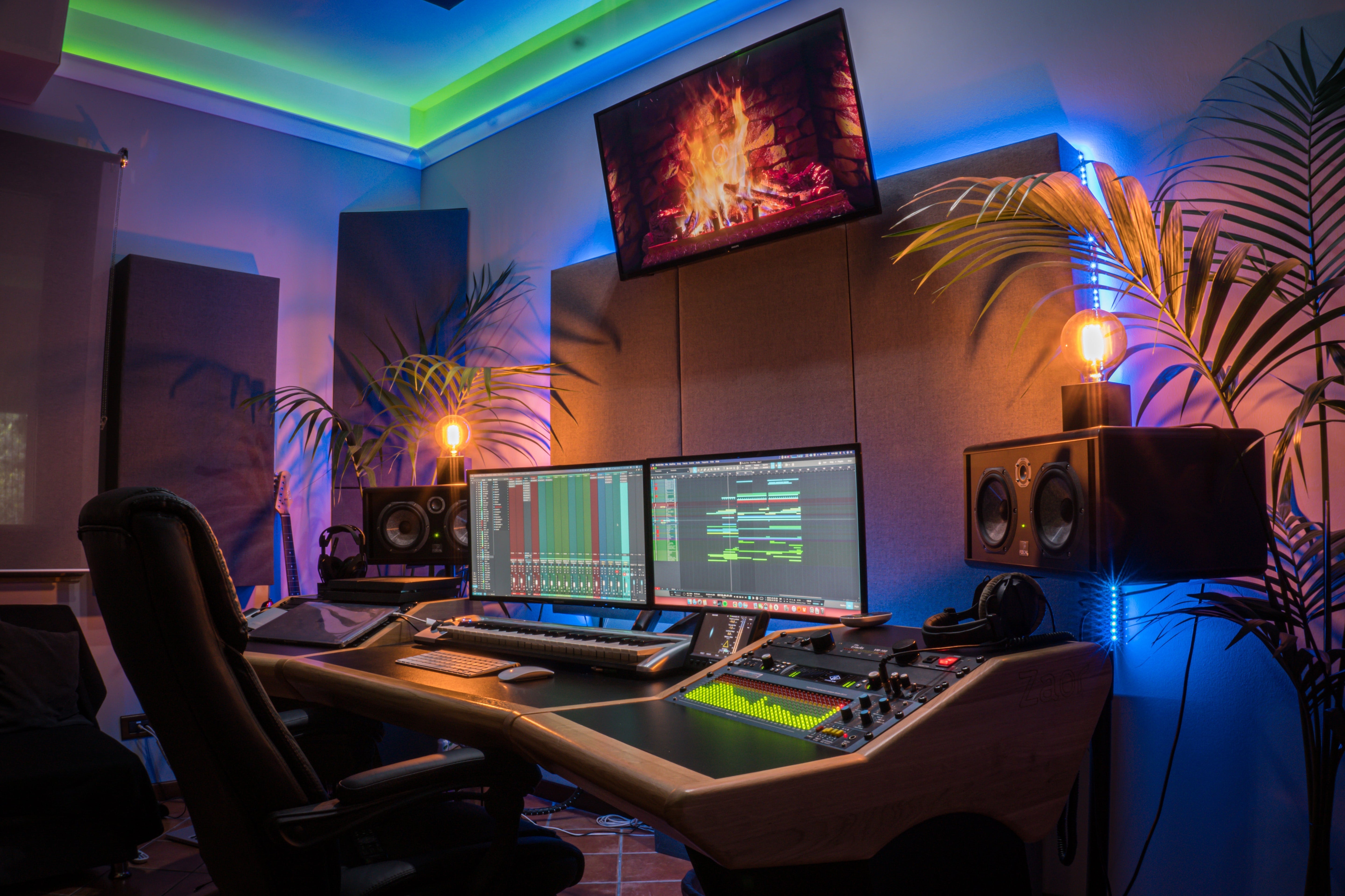 A recording studio mixing desk with dual monitors, studio monitors, and a mixing console, with GIK Acoustics acoustic panels visible on the rear wall and in the corners, lit by colourful LED lighting.